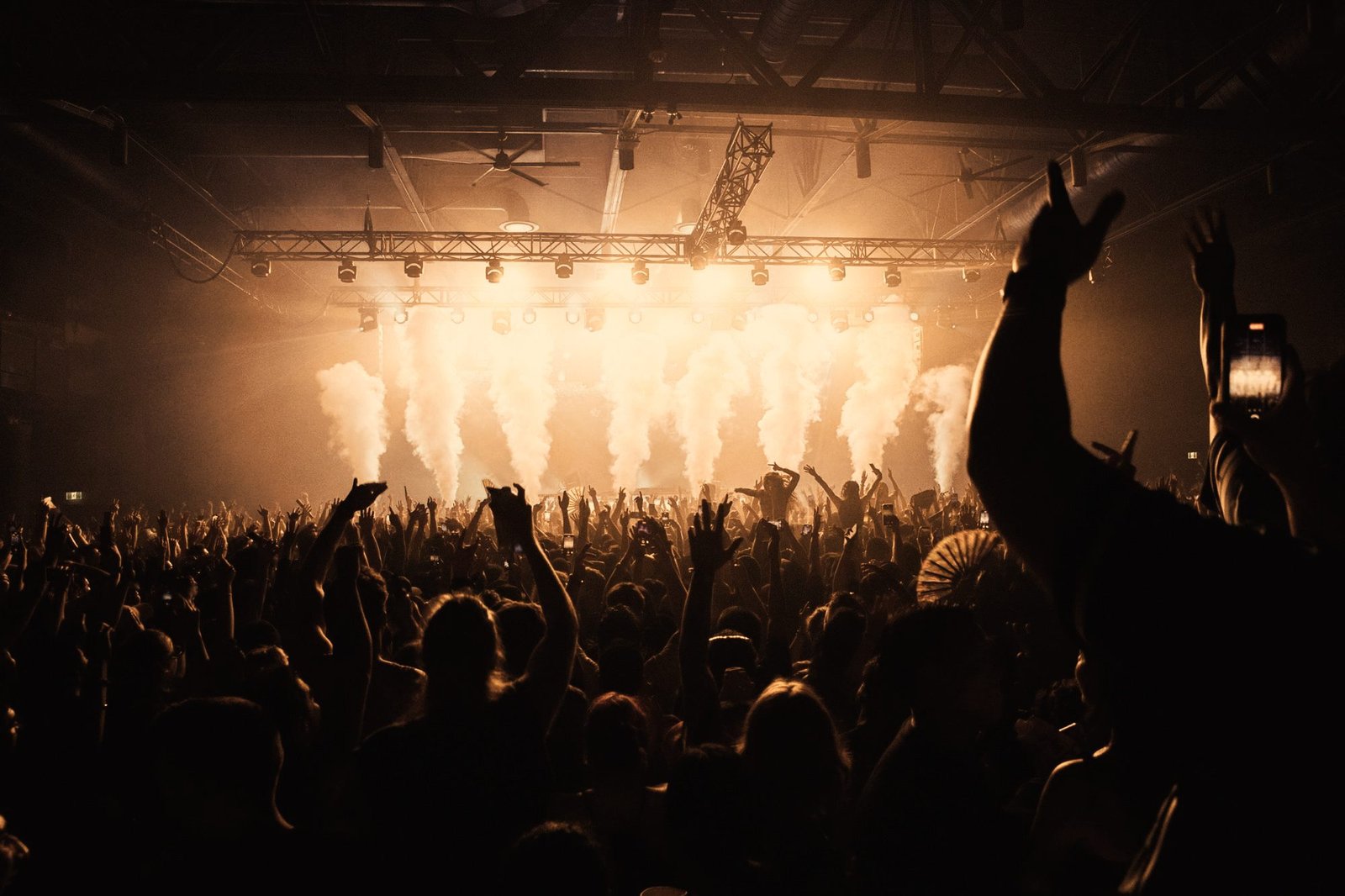 Packed crowd with hands raised in front of a lit stage firing CO2 jets